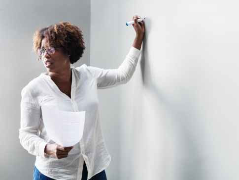 A photo of a black woman with glasses in professional attire, paper in hand, teaching while writing with a marker.